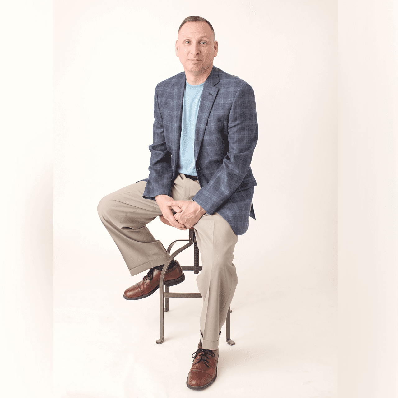 Man in a blazer sitting on a stool against a white background.